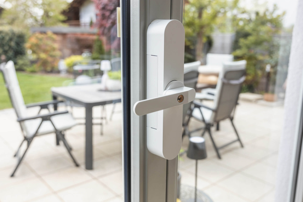 Moderne Türverriegelung an einem großen Fenster mit Blick auf eine Terrasse und Gartenmöbel.