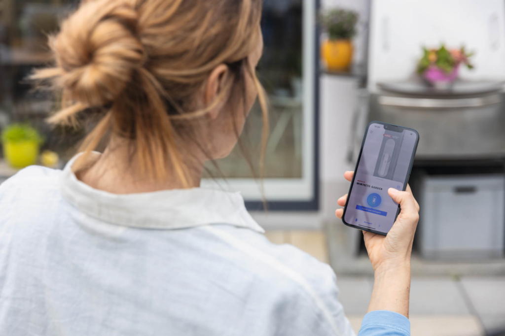 Eine Person scannt mit ihrem Smartphone ein Objekt im Freien, während sie an einem modernen Haushalt steht.